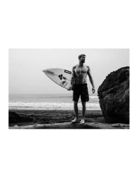 Black and white photograph of a surfer holding a surfboard standing barefoot on a sandy beach near ocean waves.