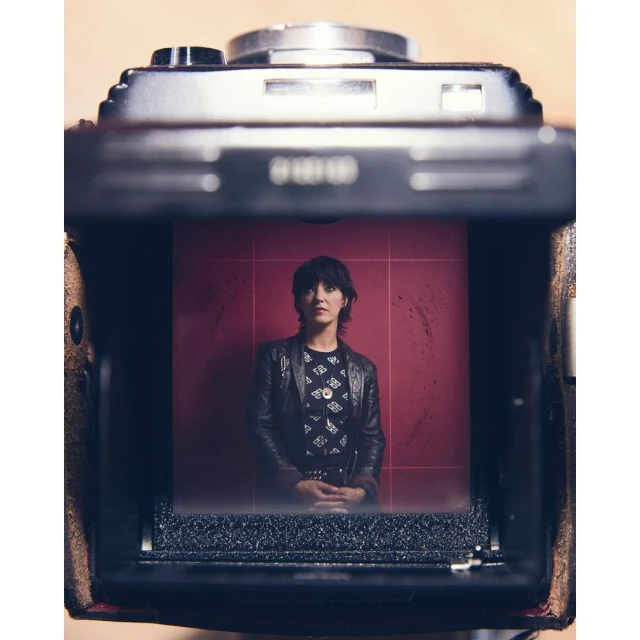 Woman with short dark hair wearing black jacket, photographed through vintage camera lens against red tile wall indoors.