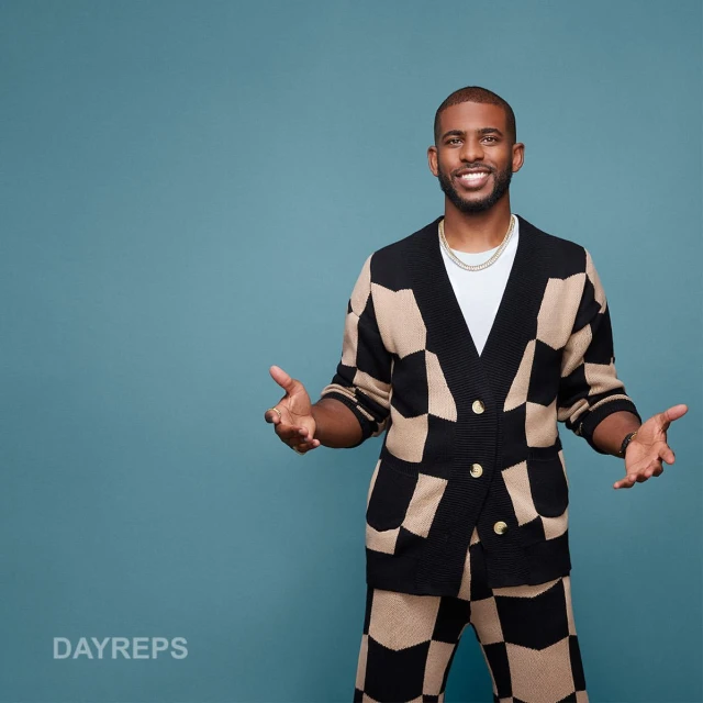 An adult male smiles wearing a checkered cardigan and pants against a blue background, with a white t-shirt and gold chain.