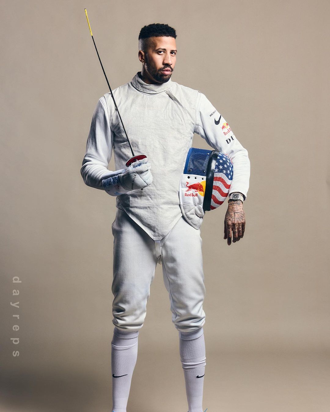 An adult male fencer in uniform poses in a studio with an epee sword and a mask adorned with the American flag.