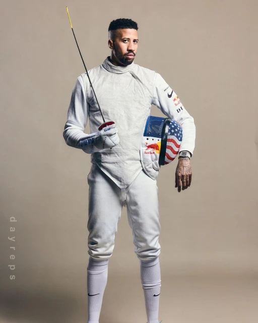 An adult male fencer in uniform poses in a studio with an epee sword and a mask adorned with the American flag.