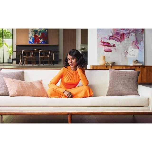 Woman in orange dress sitting cross-legged on a white couch in living room with modern art and neutral decor.