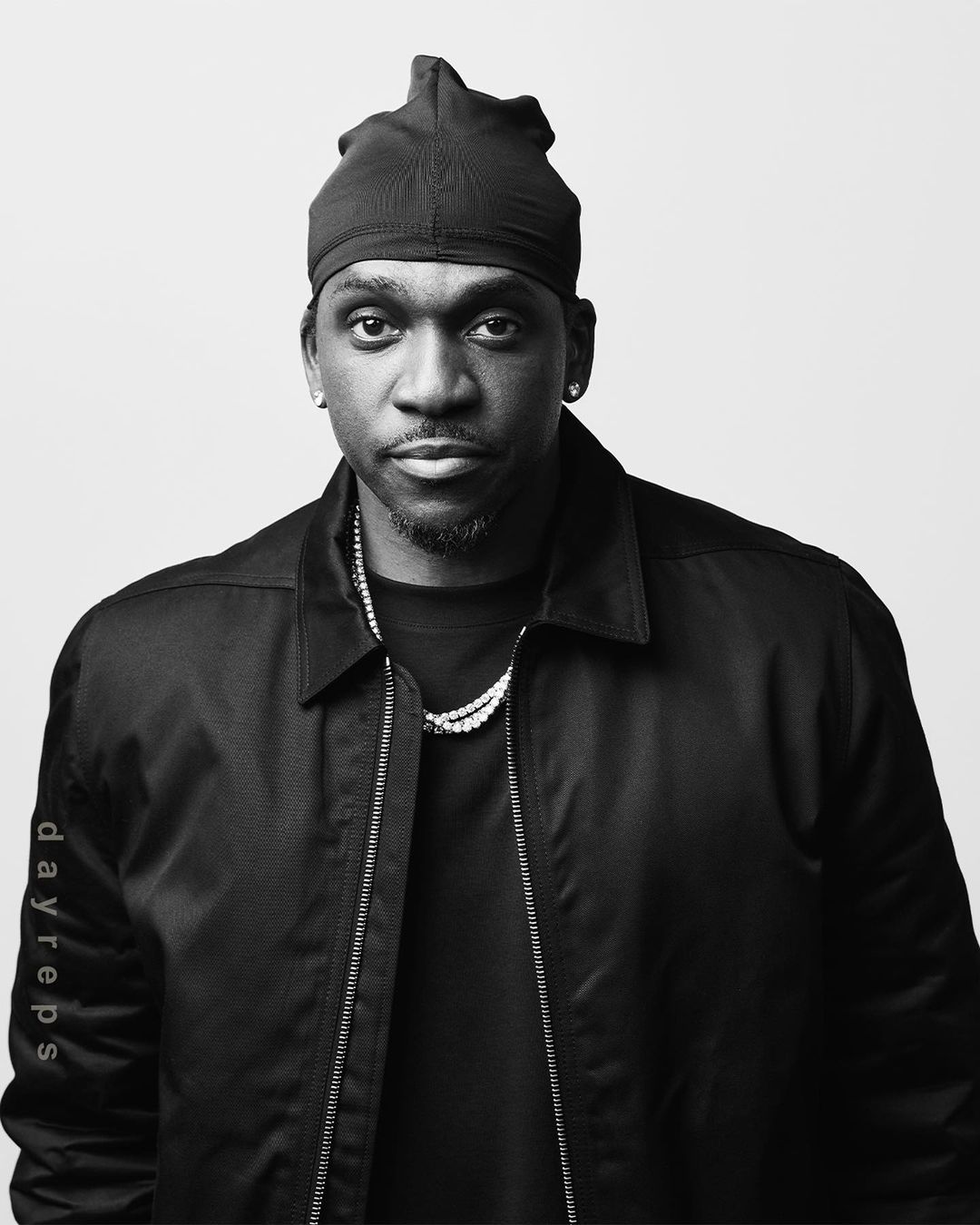 Portrait of a Black adult male wearing a black durag and jacket against a plain white backdrop, looking directly at the camera.