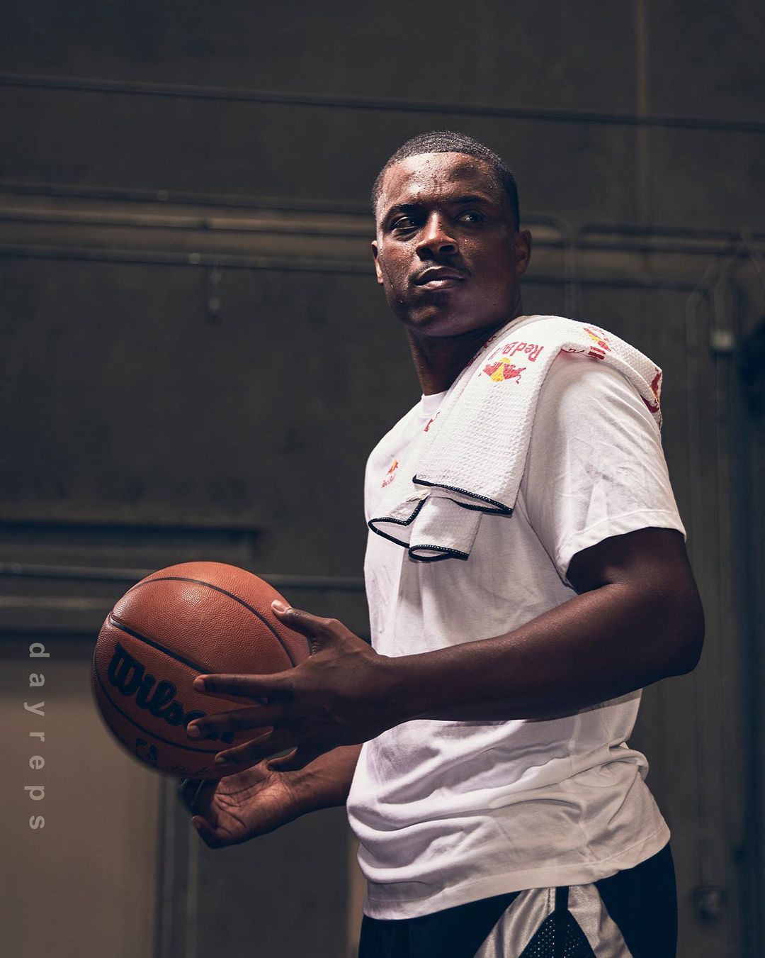 Adult man holding a Wilson basketball, wearing a white Red Bull t-shirt and white towel, in an indoor court-like setting.