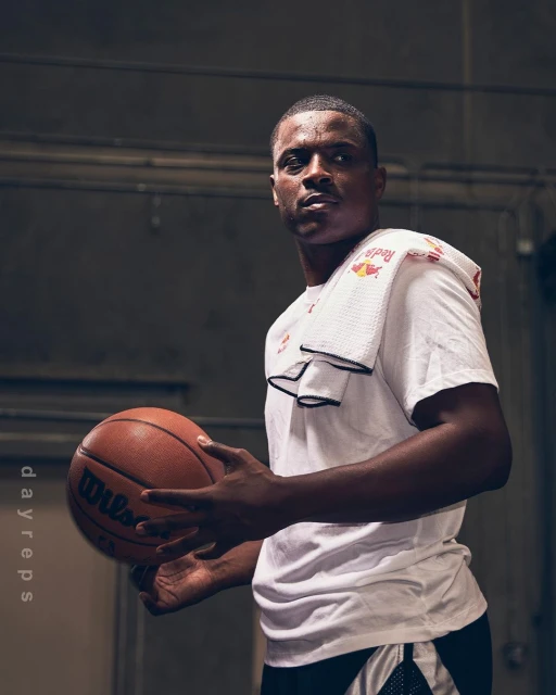 Adult man holding a Wilson basketball, wearing a white Red Bull t-shirt and white towel, in an indoor court-like setting.