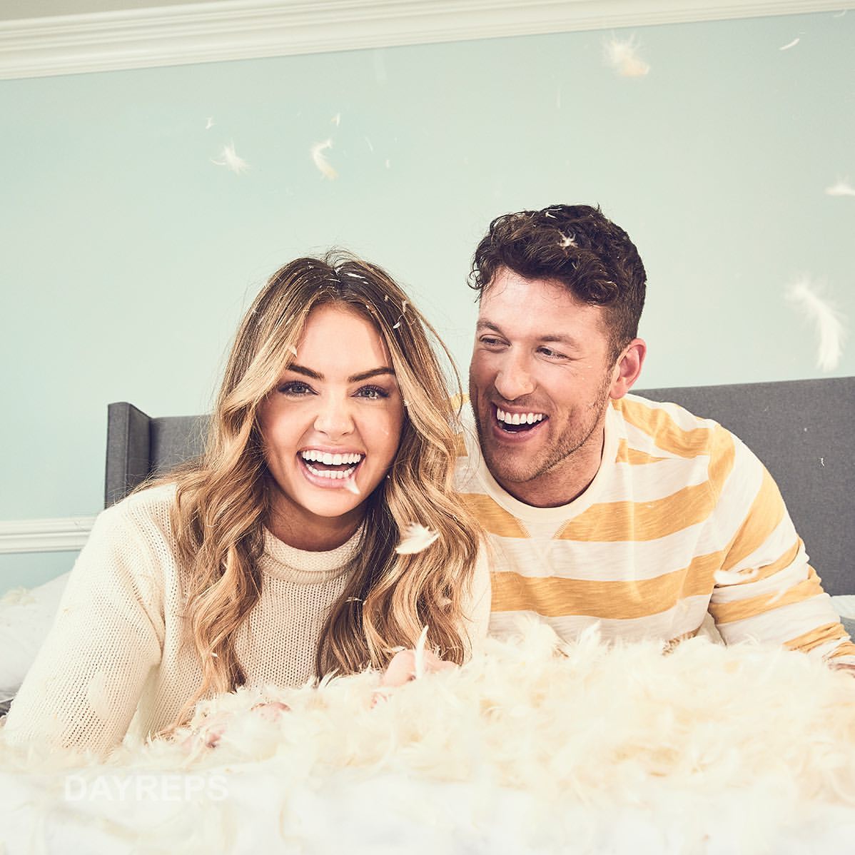 A joyful couple is lying on a bed, surrounded by feathers, both laughing with open mouths, creating a lively, lighthearted scene.