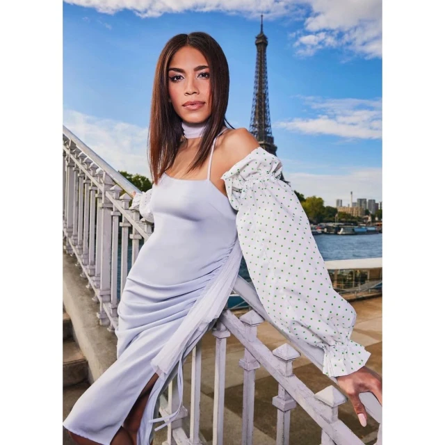 A woman poses wearing a light purple dress on a bridge with the Eiffel Tower in the background on a sunny day in an outdoor shot.