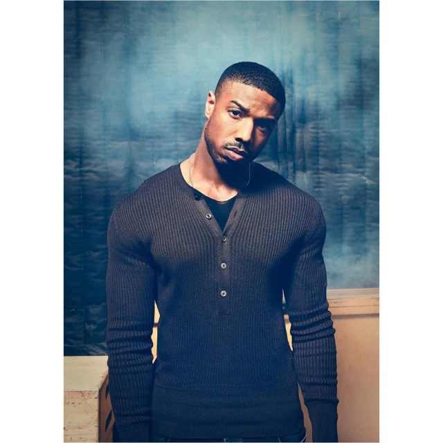 Portrait of a handsome African-American man with short hair wearing a dark ribbed henley shirt, set against a textured blue background.
