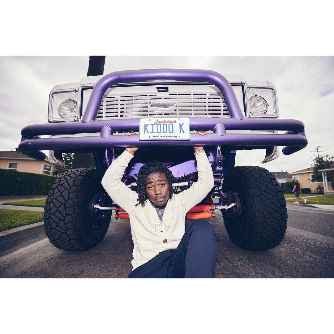 A man with dreadlocks sits beneath a purple truck with oversized tires on a suburban street, holding the license plate.