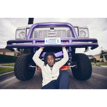 A man with dreadlocks sits beneath a purple truck with oversized tires on a suburban street, holding the license plate.
