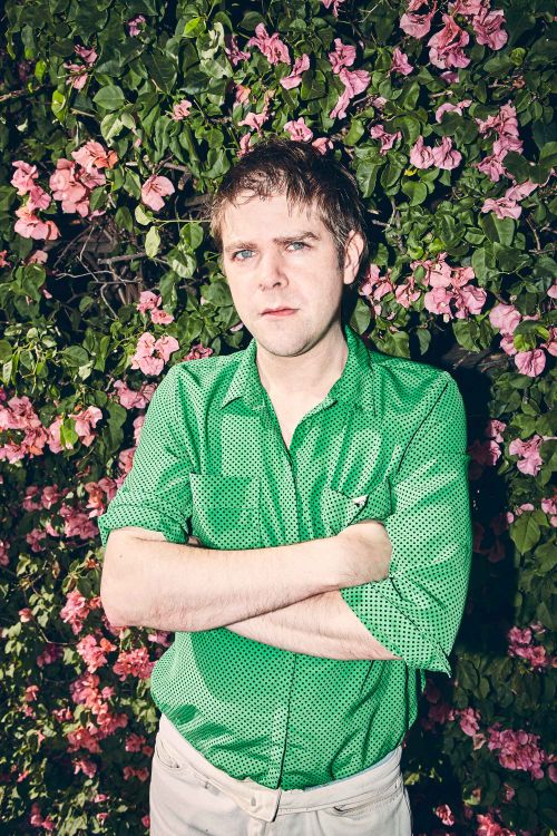 Portrait of a man in a green shirt with arms crossed, standing in front of a blooming bougainvillea bush with pink flowers.