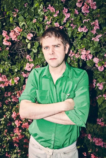 Portrait of a man in a green shirt with arms crossed, standing in front of a blooming bougainvillea bush with pink flowers.