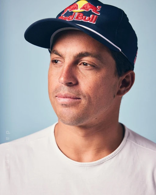 Portrait of a man wearing a Red Bull baseball cap against a light blue background, looking off to the side with a slight smile.