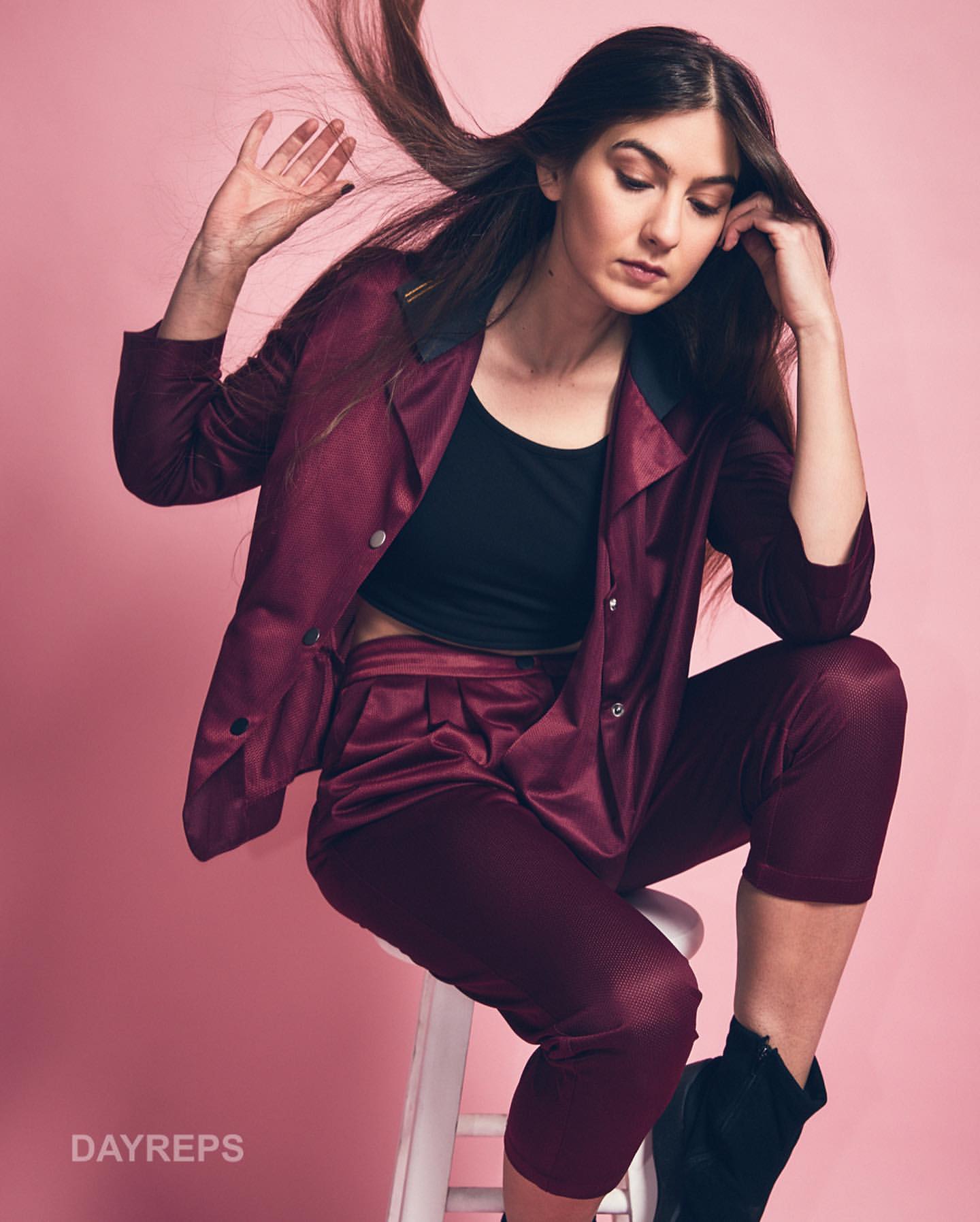 Portrait of a woman wearing a maroon pantsuit and black crop top seated on a white stool against a pink background
