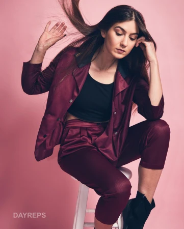 Portrait of a woman wearing a maroon pantsuit and black crop top seated on a white stool against a pink background