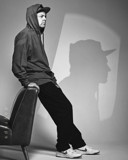 Black and white portrait of a man with a baseball cap and hoodie, sitting on an arm chair with a shadow cast on the wall behind him.