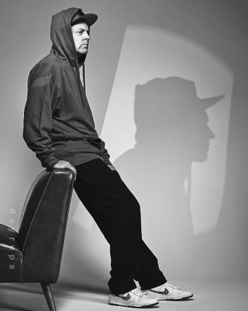 Black and white portrait of a man with a baseball cap and hoodie, sitting on an arm chair with a shadow cast on the wall behind him.