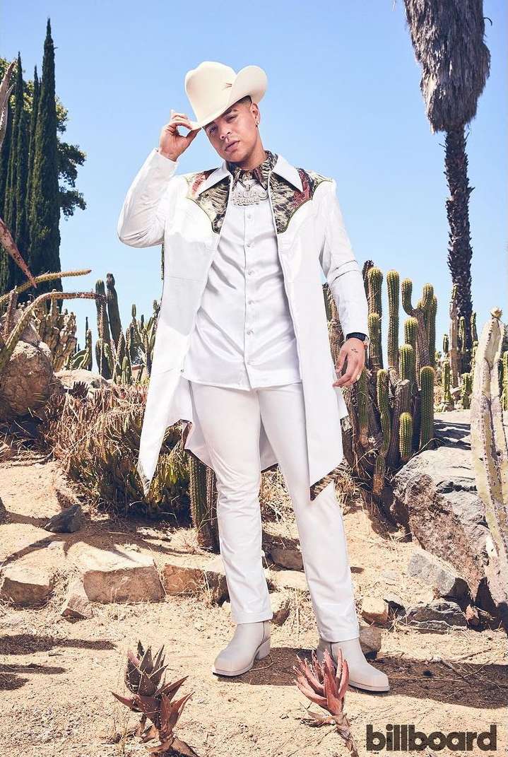 Man in white cowboy hat, coat, and pants poses in arid landscape with cacti and blue sky for fashion, music, or entertainment visual content.