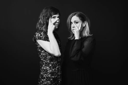 Black and white portrait of two adult women in dark dresses posing with hands near their faces against a dark backdrop in studio.