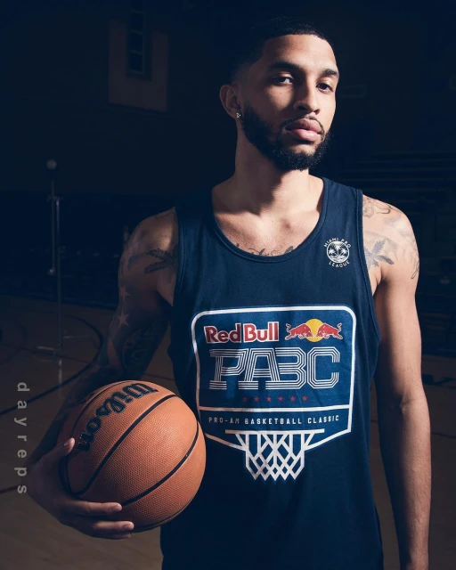 Adult male in a tank top holds a Wilson basketball in an indoor setting. The tank has a Red Bull PABC logo.