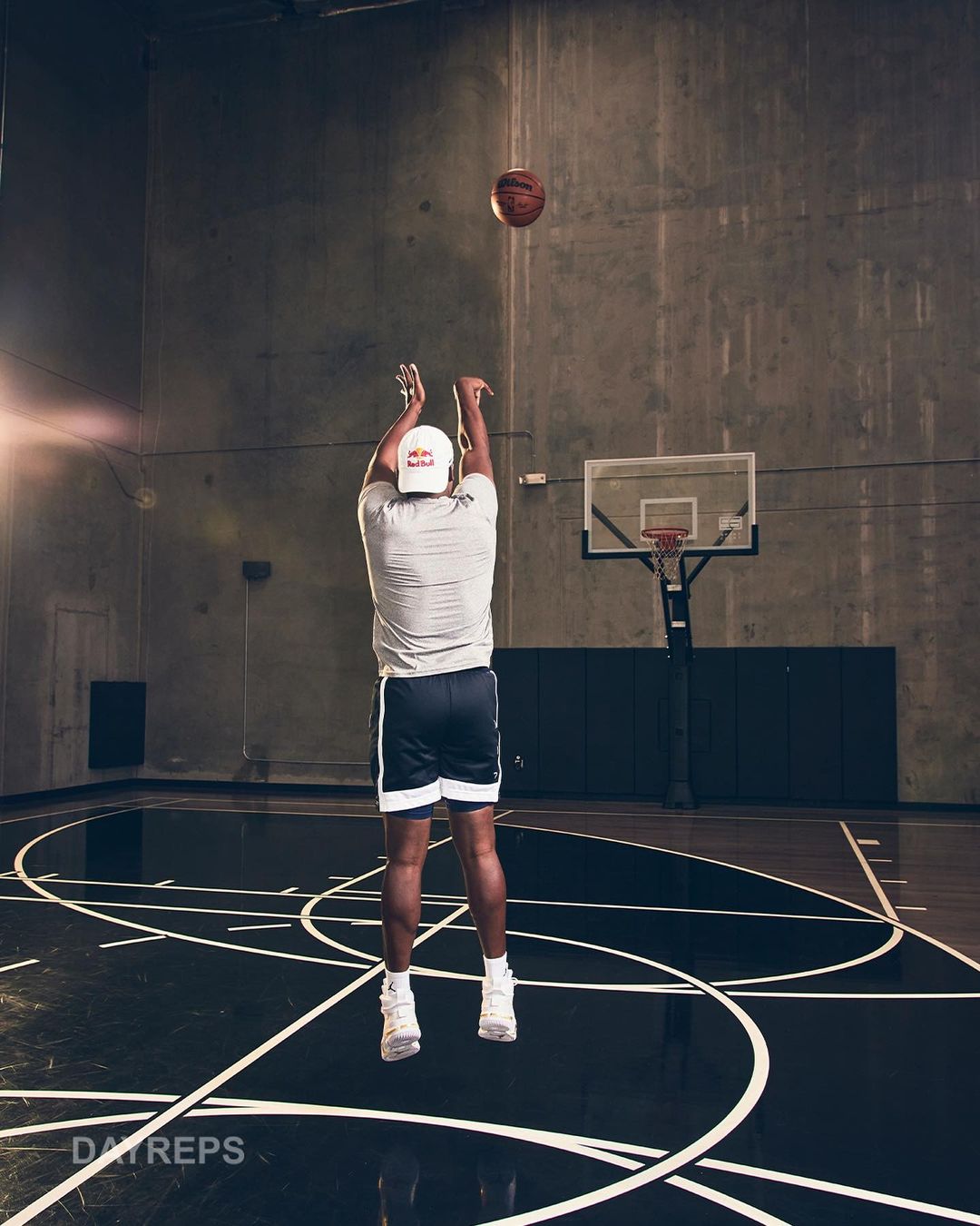 Basketball player in athletic shorts and a tee-shirt, suspended mid-air, shooting a basketball towards a hoop inside a basketball court.
