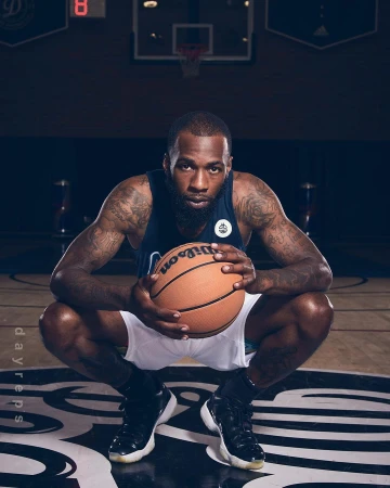 Bearded African-American man in athletic wear squats with Wilson basketball in indoor sports setting with court markings visible.