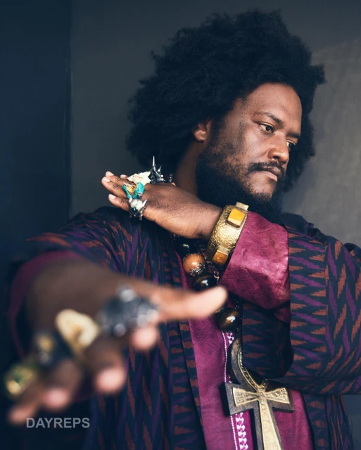 Portrait of adult male with dark skin and afro hair wearing ethnic clothing and jewelry, with a blurred hand in the foreground.