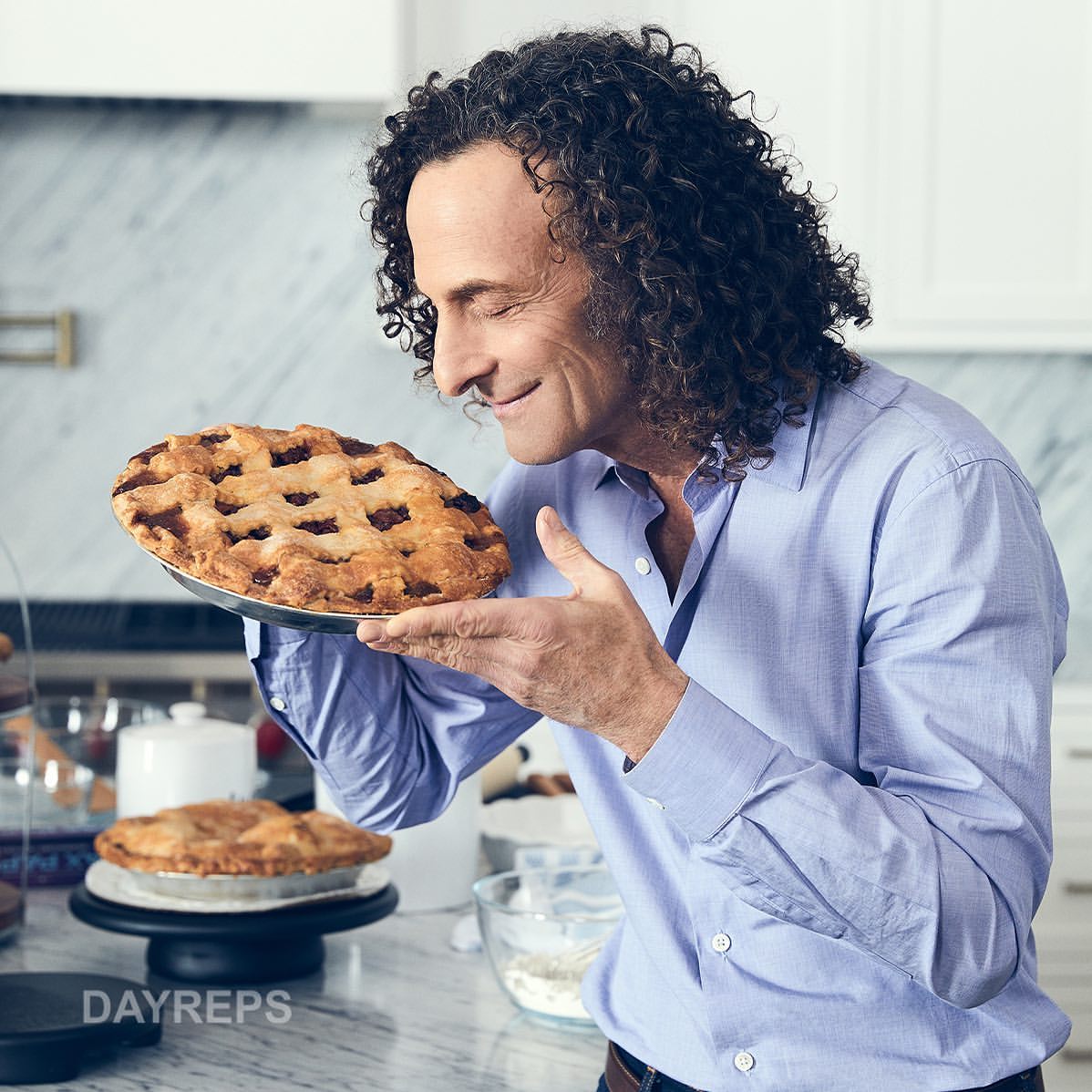 Adult male in a blue shirt smiles while holding a lattice-crust pie in a kitchen with a white marble countertop and cabinets.