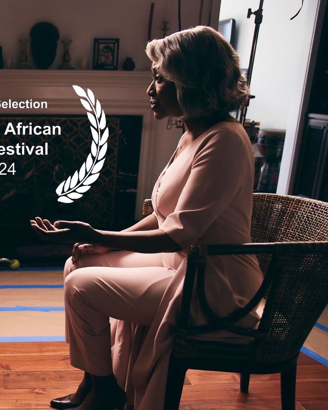 Woman in a light pink jumpsuit sits in a wicker chair indoors, with an award graphic and film festival text overlay.