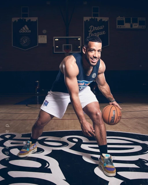 Adult male basketball player dribbling Wilson basketball on indoor court with Adidas and Drew League banners hanging in the background.