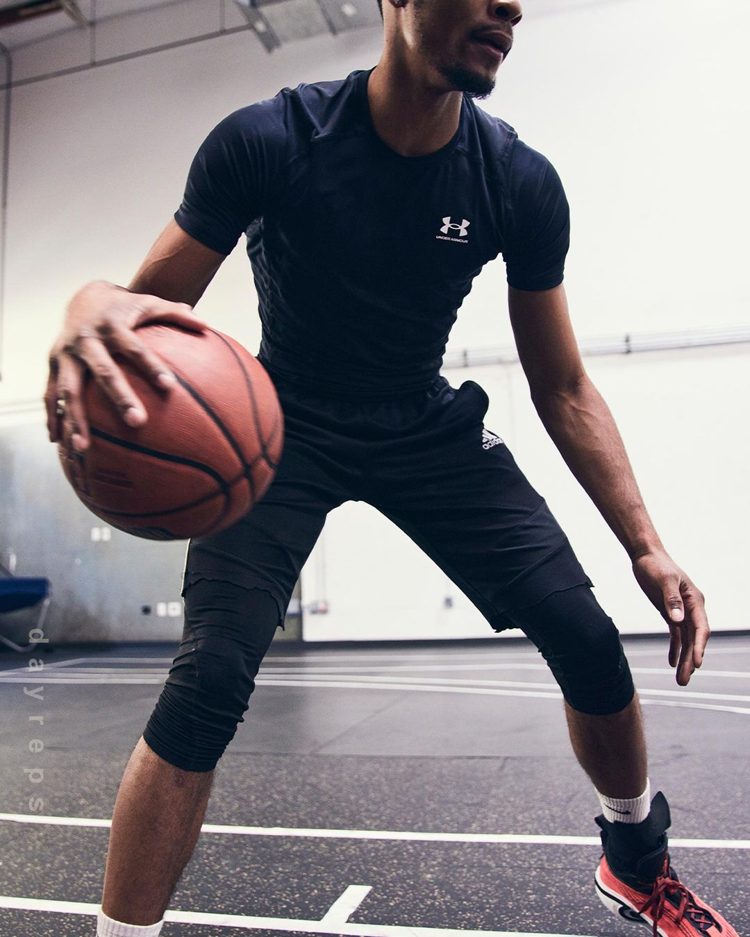 An athletic man dribbles a basketball on an indoor court, wearing a dark T-shirt, black shorts, and red and black athletic shoes.