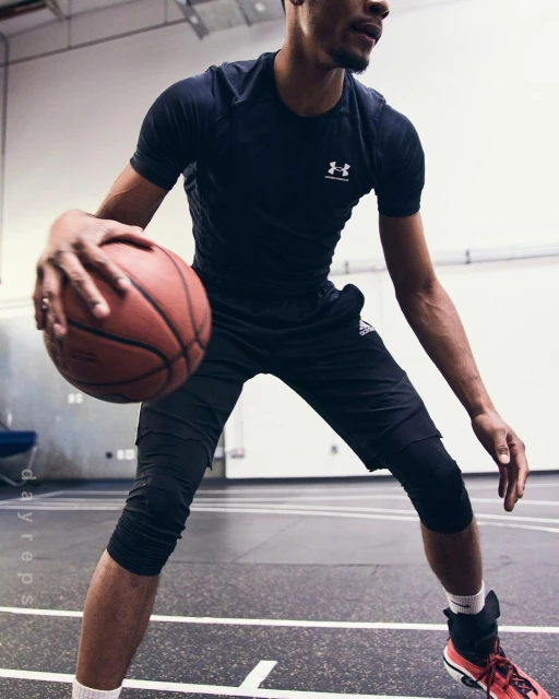An athletic man dribbles a basketball on an indoor court, wearing a dark T-shirt, black shorts, and red and black athletic shoes.