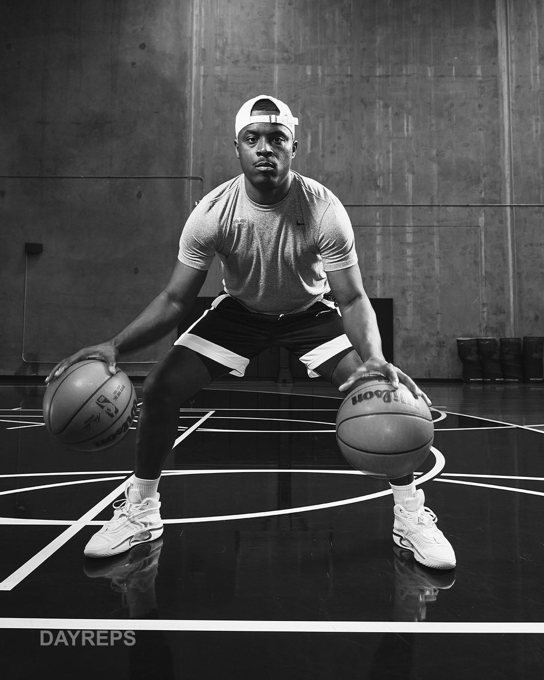 A Black man dribbles two basketballs indoors, wearing a hat, t-shirt, and shorts, with a concrete wall behind and lines on the floor.