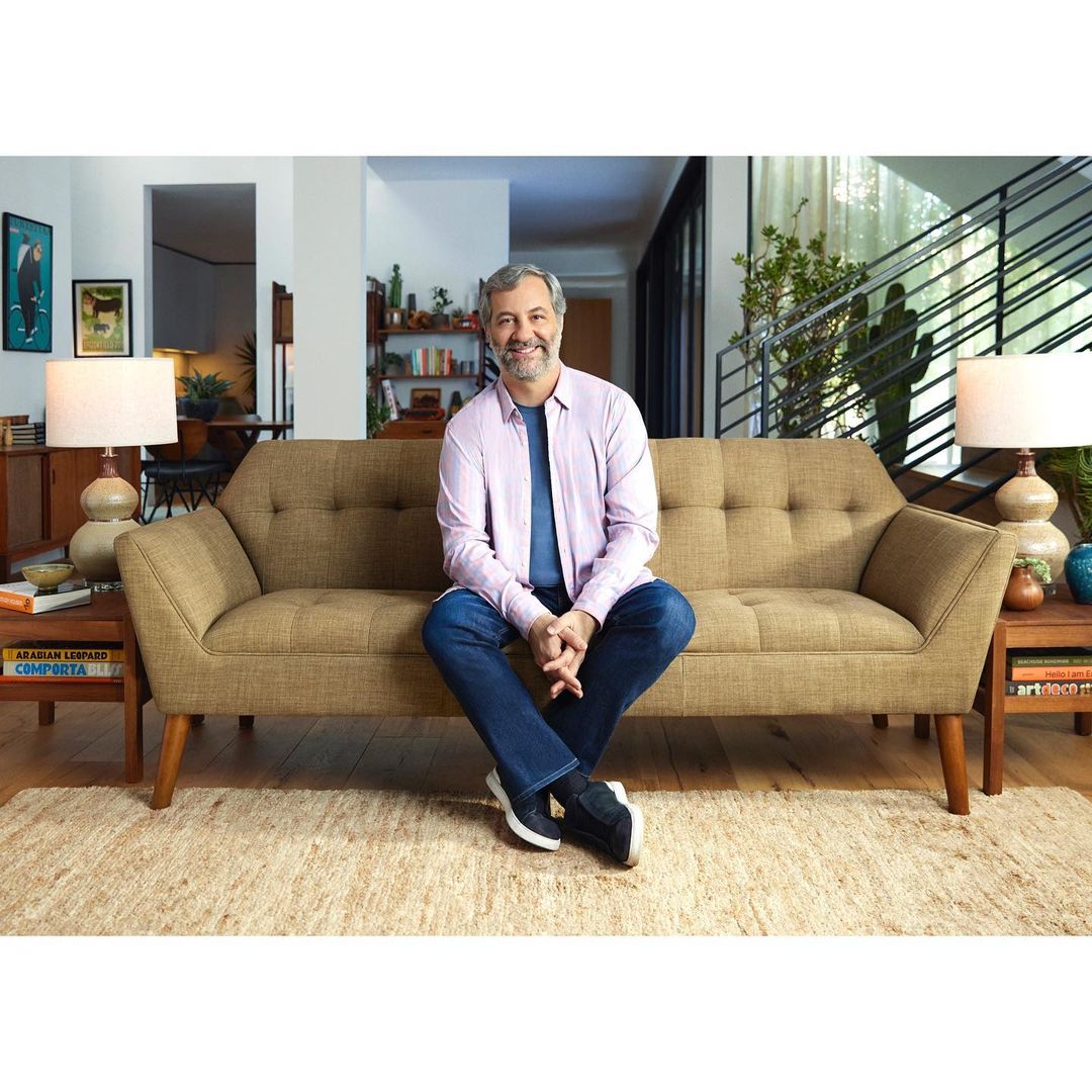 Adult male sitting on a tan sofa in a well-lit living room with neutral tones and modern decor, creating a comfortable and inviting atmosphere.