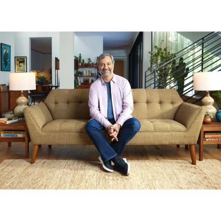 Adult male sitting on a tan sofa in a well-lit living room with neutral tones and modern decor, creating a comfortable and inviting atmosphere.