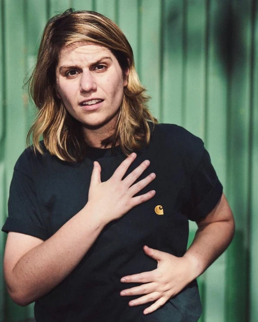 Blonde-haired adult female in a dark-green Carhartt t-shirt with hand on her chest, posing indoors against a green background.