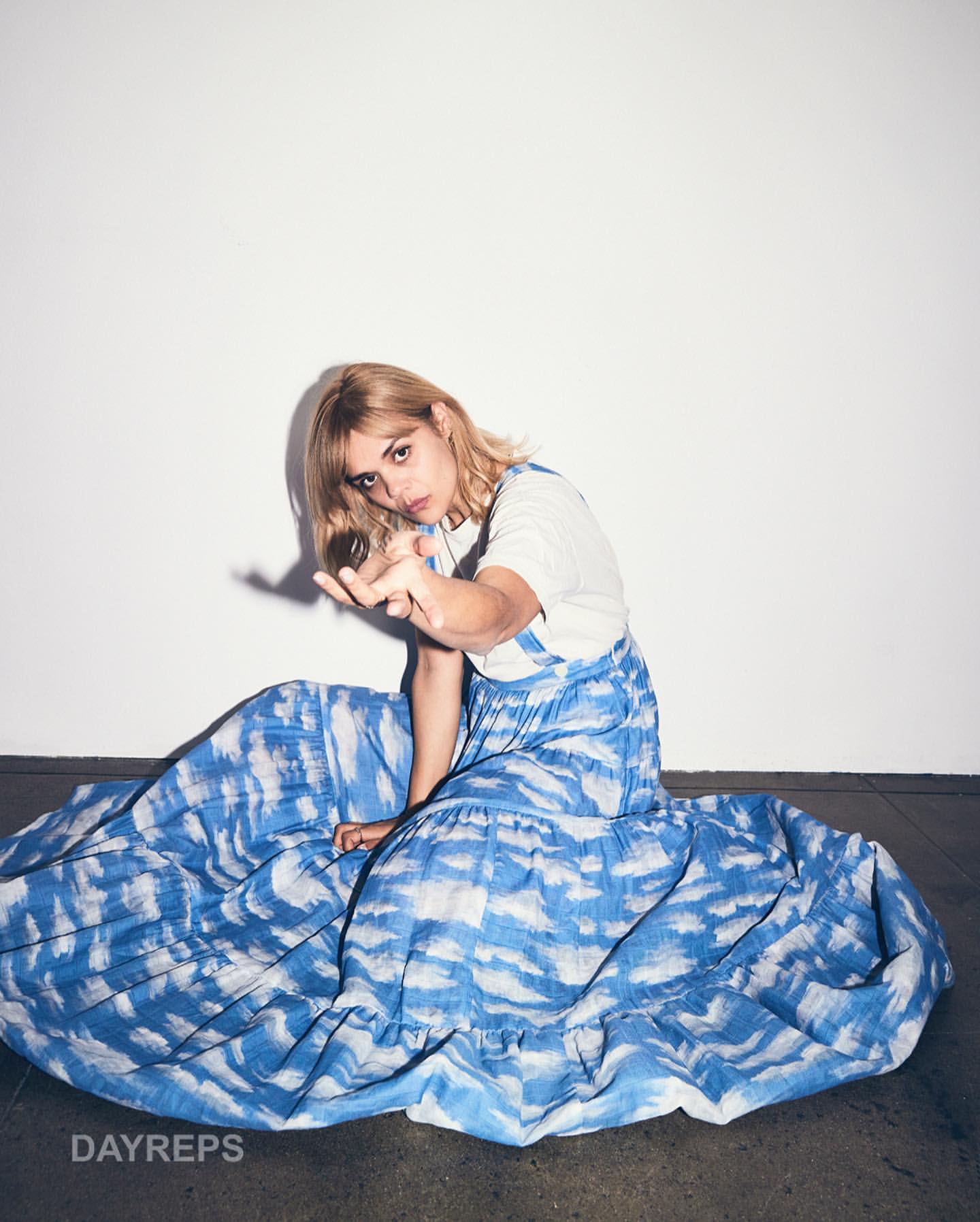 Blonde woman in a tie-dye maxi dress sitting indoors with arm extended toward the camera against a stark white background.