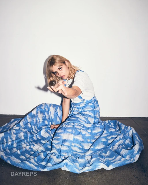 Blonde woman in a tie-dye maxi dress sitting indoors with arm extended toward the camera against a stark white background.