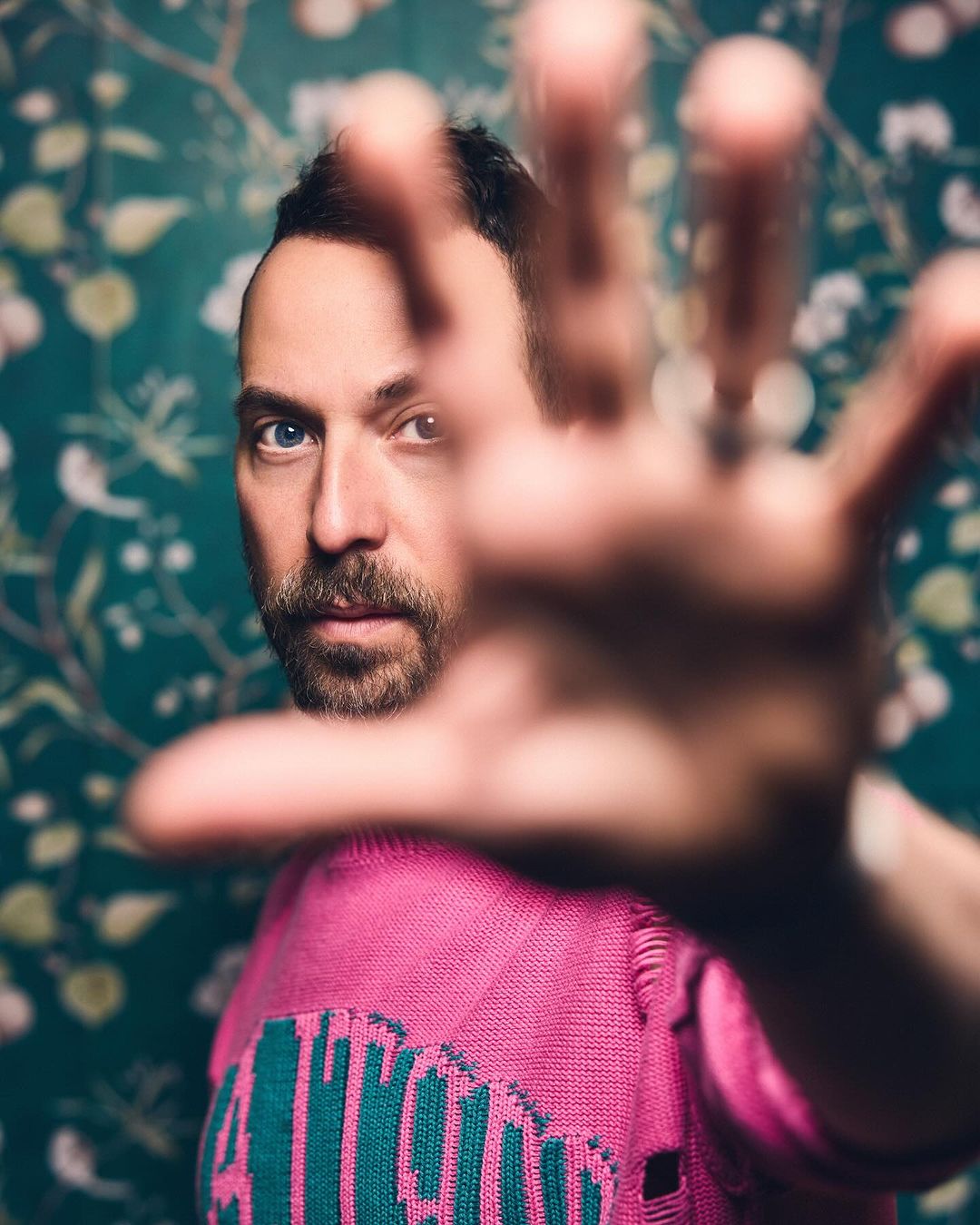 Portrait of an adult male with a beard wearing a pink sweater, with a hand reaching out toward the camera against a floral wallpaper.