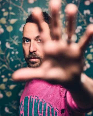 Portrait of an adult male with a beard wearing a pink sweater, with a hand reaching out toward the camera against a floral wallpaper.
