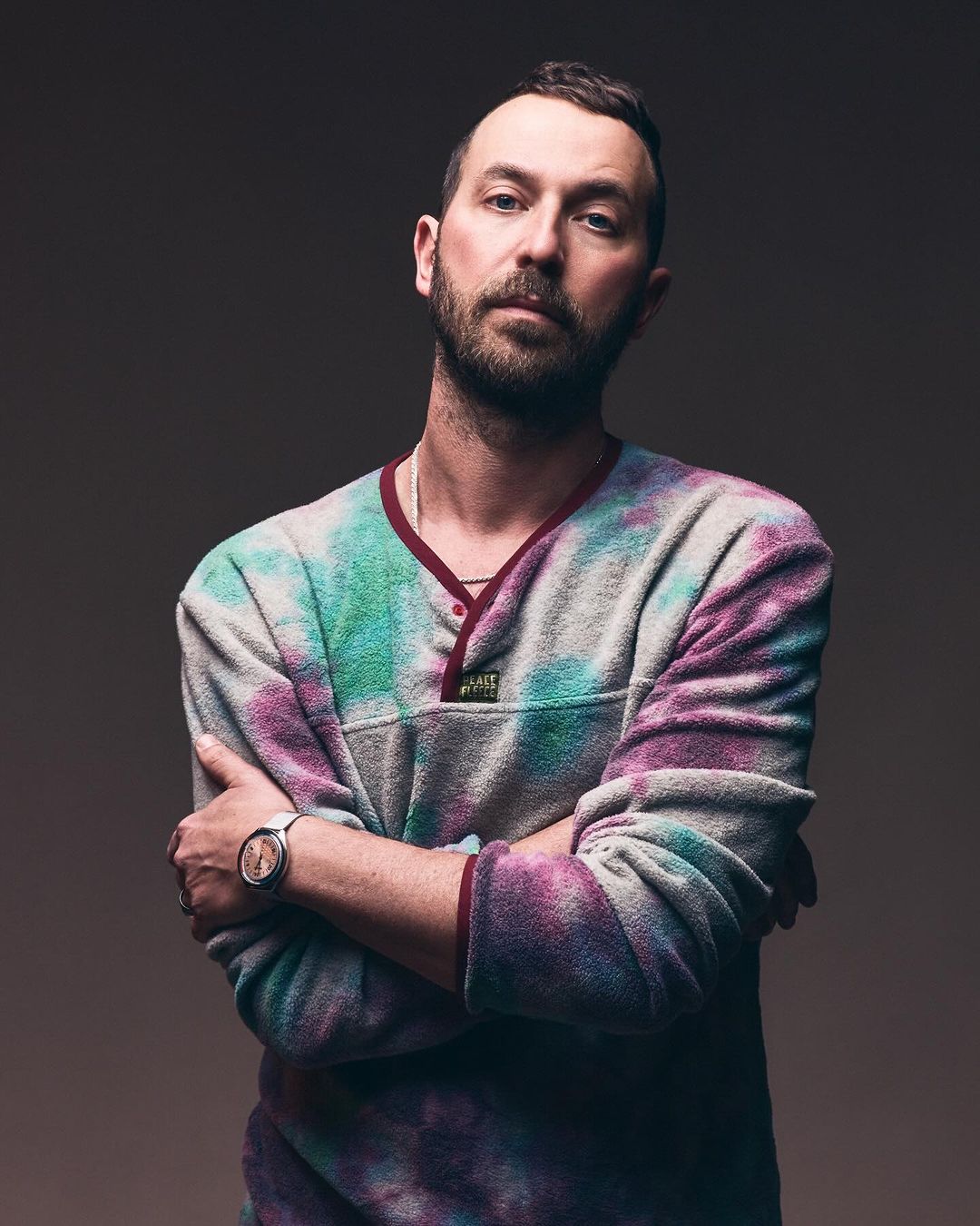 Portrait of adult male with crossed arms in tie-dye fleece pullover, wearing a watch, against a neutral gradient background.