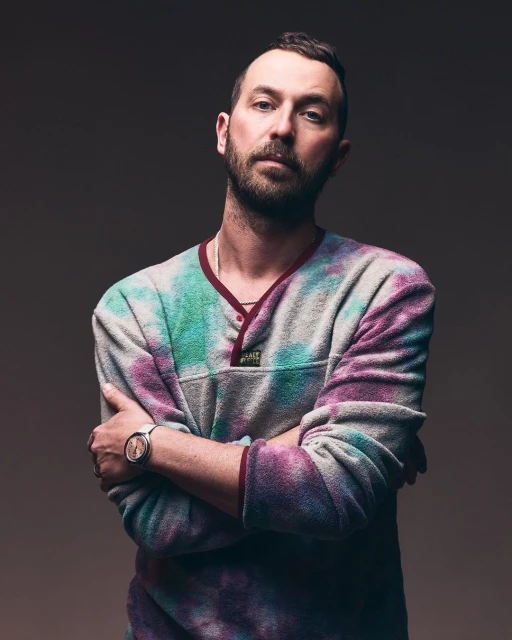 Portrait of adult male with crossed arms in tie-dye fleece pullover, wearing a watch, against a neutral gradient background.