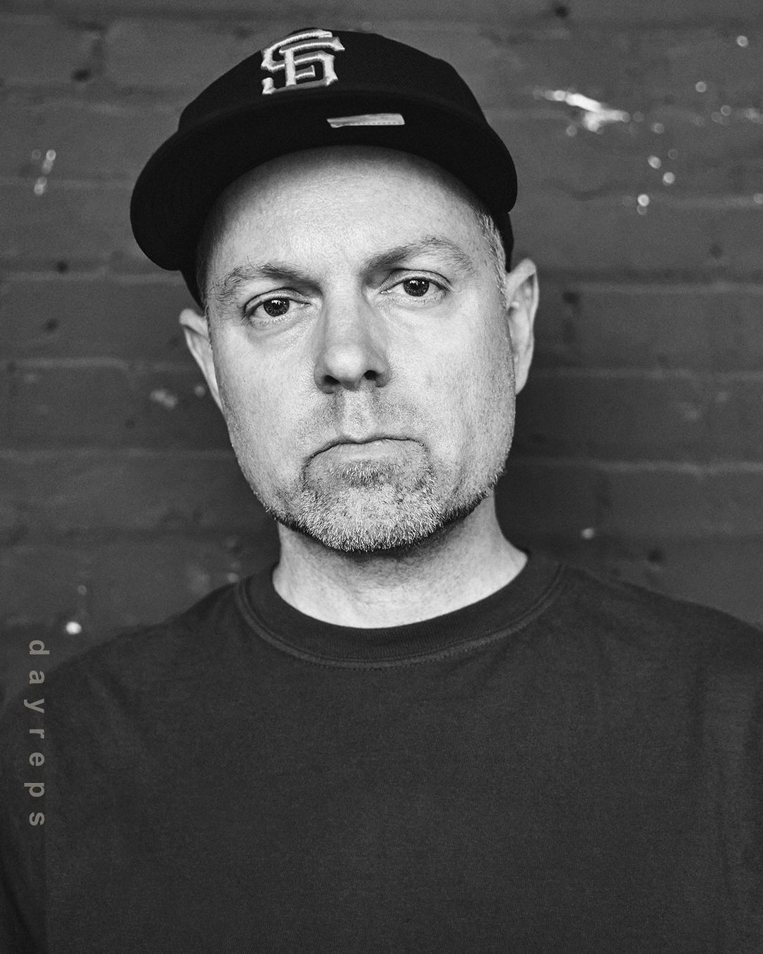 Headshot of an adult male wearing a baseball cap with the SF logo, black t-shirt, and a short beard, posing indoors against a brick wall.