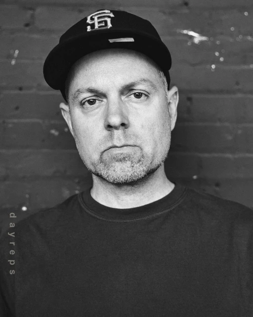 Headshot of an adult male wearing a baseball cap with the SF logo, black t-shirt, and a short beard, posing indoors against a brick wall.