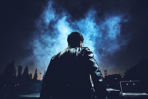 Man with back to camera in leather jacket with smoke effect, stage lights, and laptop in a night scene. Music industry vibe.