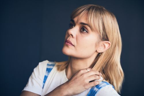 Portrait of a blond-haired woman in a white t-shirt looking upwards against a dark background in an indoor environment, medium shot.