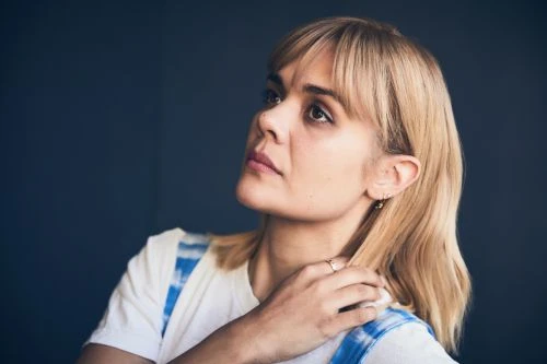 Portrait of a blond-haired woman in a white t-shirt looking upwards against a dark background in an indoor environment, medium shot.
