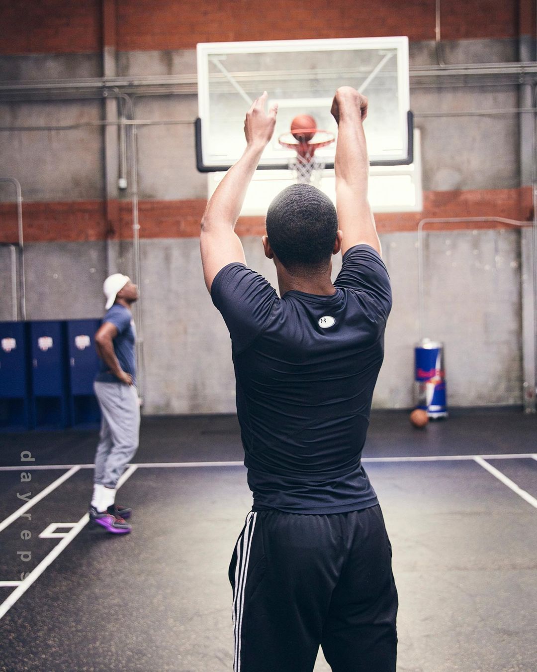 A man wearing athletic clothing throws a basketball at an indoor gym, as another man stands in the background with a Red Bull can visible.