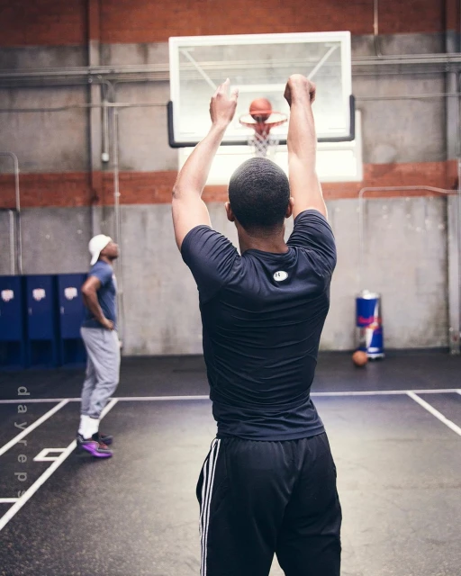 A man wearing athletic clothing throws a basketball at an indoor gym, as another man stands in the background with a Red Bull can visible.