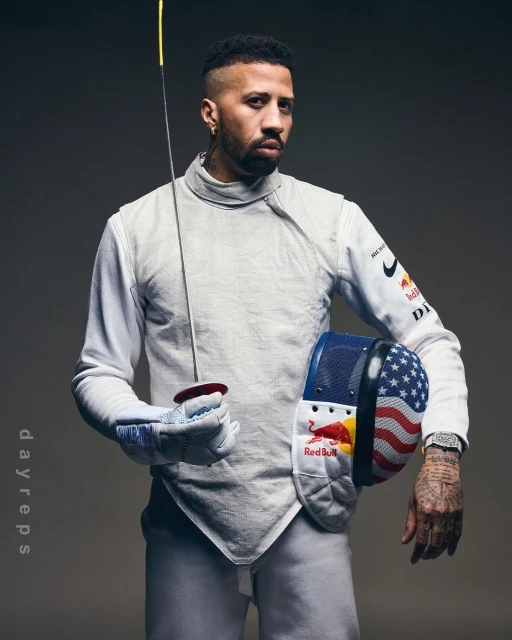 Portrait of a male fencer in fencing uniform holding a foil and fencing mask decorated with the American flag and Red Bull logos.